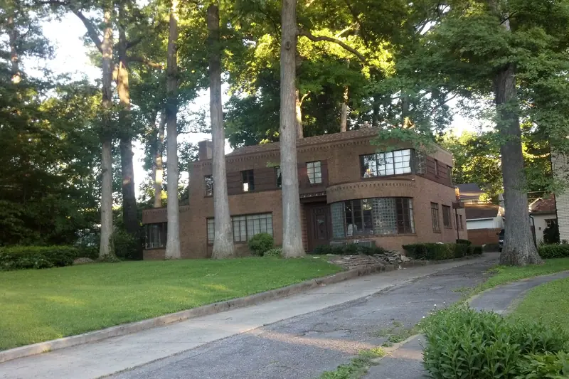 Brown brick two-story house with glass-block basement windows, set back among big tree trunks on a sloping yard