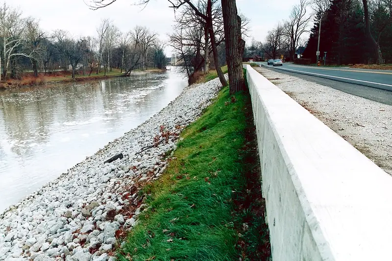 Concrete flood wall topped with a rail and backed by rock armor along the St. Joseph River, bare trees on the far bank