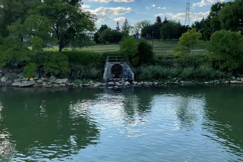 Rectangular concrete outfall opening in a rock-lined riverbank across a calm stretch of green river